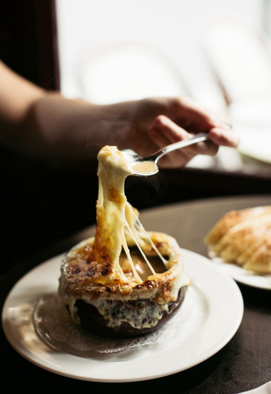 Person dipping a spoon into a rich cheese fondue served in a bread bowl on a white plate.