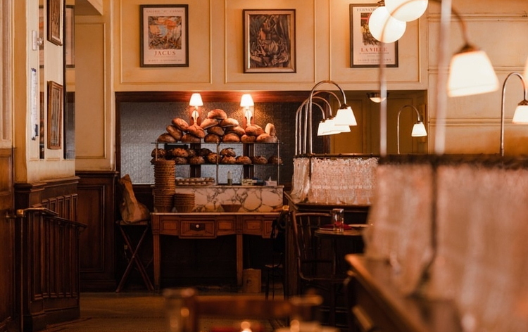 Warm indoor setting with wooden furniture, bread display, and hanging pendant lights in a cozy restaurant atmosphere.