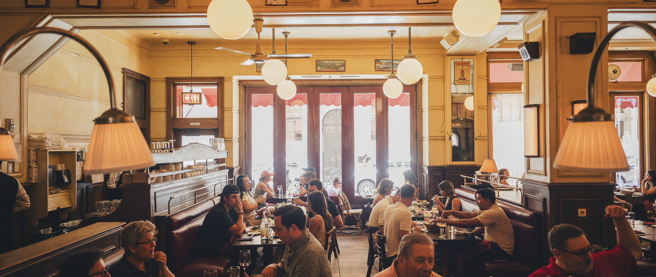 Vintage restaurant interior with patrons dining at tables, lace curtains, warm lighting, and large windows.