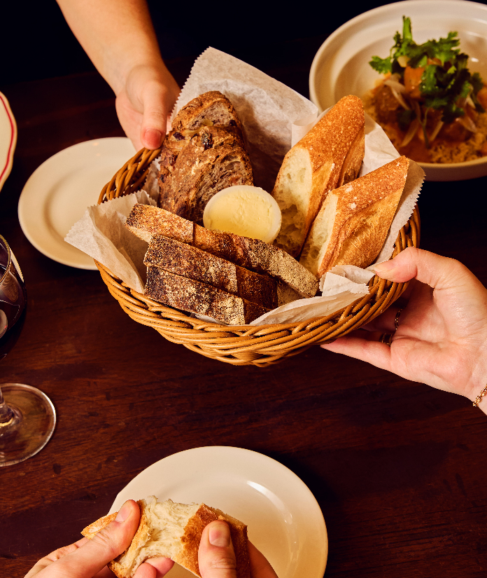 Two people passing a woven bread basket filled with sliced baguette, dark bread, and a pat of butter over a dark wooden restaurant table, with small plates, a wine glass, and a dish with herbs visible in the background.