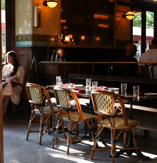A sunlit French bistro dining area with woven rattan chairs, a dark wooden banquette, and neatly set tables with glassware, as a woman stands near the entrance in the background.