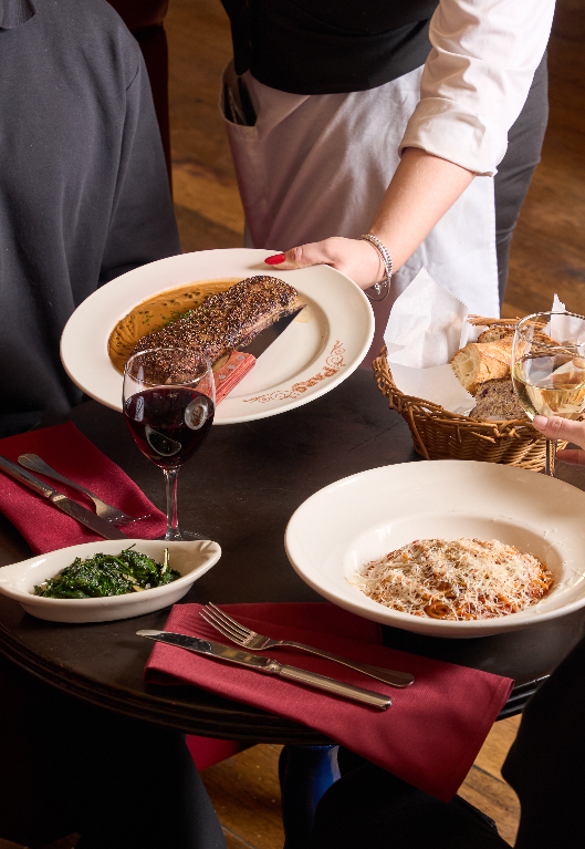 A server placing a plated steak at a restaurant table set with a glass of red wine, a pasta dish, a side of sautéed greens, a bread basket, and deep red linen napkins.
