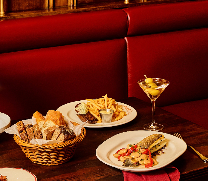 Restaurant table spread with burger and fries, bread basket, fish dish with vegetables, and martini against red booth seating